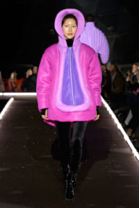Runway model wearing a bright pink oversized hooded coat with a purple front panel, black pants, and glossy black boots.