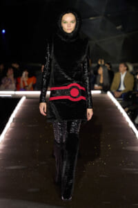 Model walking runway in black fur outfit with a wide red belt and hooded top, audience in background.