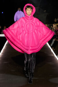 Model walking a runway in a bright pink oversized padded cape with a hood.