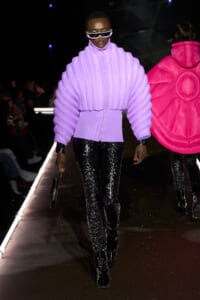 Model walks the runway wearing a lavender oversized quilted jacket, white futuristic sunglasses, and glossy black pants.