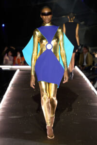Model walks a runway in a futuristic metallic gold bodysuit with a royal blue geometric overlay and cyan cape, wearing a dark visor.