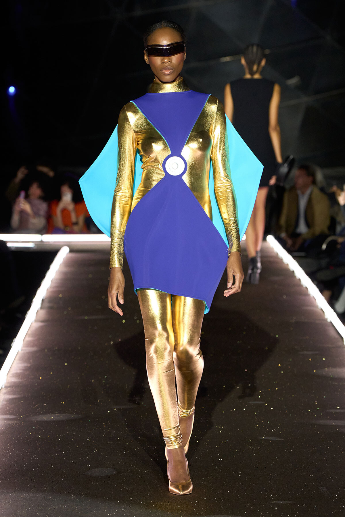 Model walks a runway in a futuristic metallic gold bodysuit with a royal blue geometric overlay and cyan cape, wearing a dark visor.