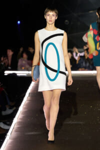 Model walking the runway in a white sleeveless shift dress with black and teal abstract curves, holding a light blue clutch.