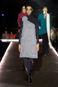 Model on a runway wearing a gray sleeveless dress with large circular appliques, black turtleneck sleeves, and oversized sunglasses.
