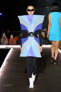 Model on a runway wearing a bold sculptural blue and purple geometric dress with black sleeves, a metallic belt centerpiece, oversized sunglasses, and white ankle boots.