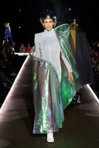 Model walking a fashion show in a silver-gray zippered jumpsuit with an iridescent metallic cape and a black helmet.