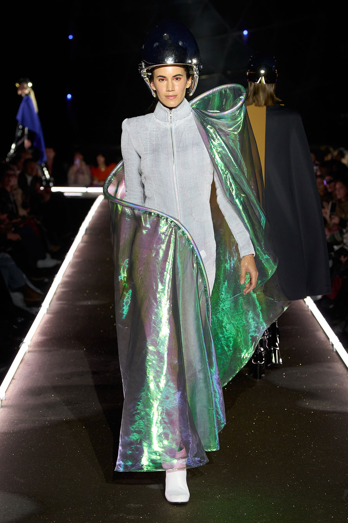 Model walking a fashion show in a silver-gray zippered jumpsuit with an iridescent metallic cape and a black helmet.