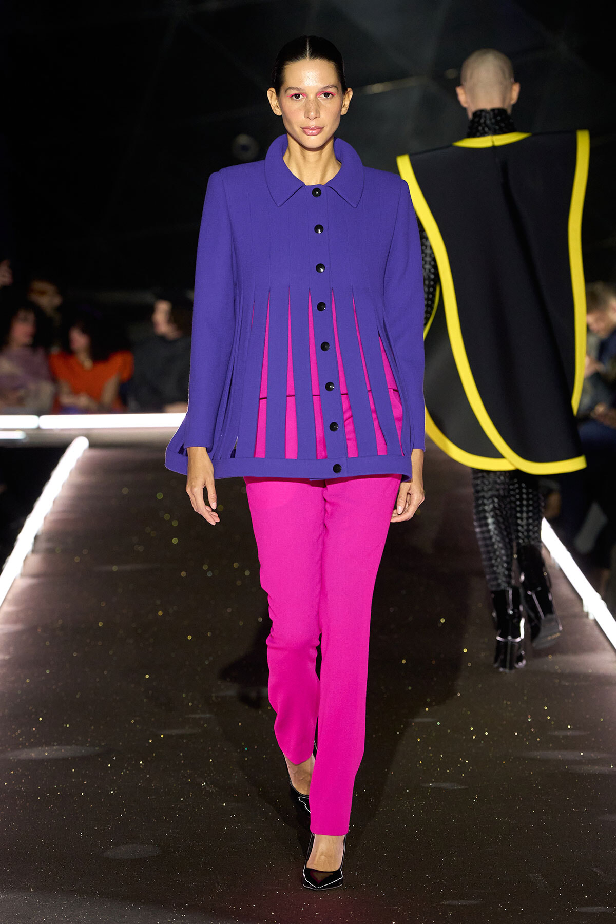 Model walks the runway wearing a royal purple coat with magenta-pink pants and black heels.",