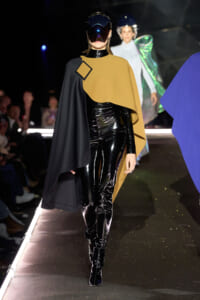 Model walks the runway in a black patent bodysuit with a tan cape draped over one shoulder and a futuristic visor helmet.