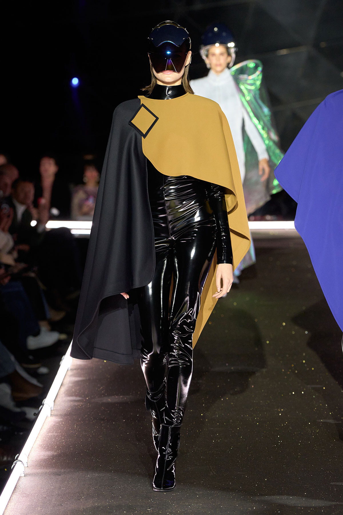 Model walks the runway in a black patent bodysuit with a tan cape draped over one shoulder and a futuristic visor helmet.