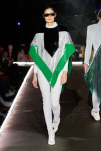Model walking a runway in a futuristic silver, black, and green angular poncho with white leggings and white boots; wearing oversized visor sunglasses.