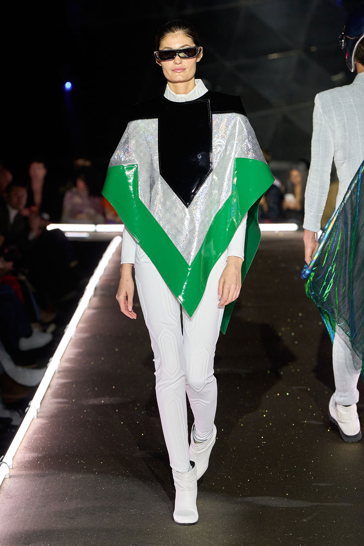 Model walking a runway in a futuristic silver, black, and green angular poncho with white leggings and white boots; wearing oversized visor sunglasses.