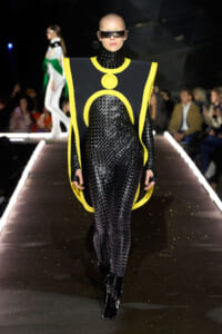 Model walks the runway in a black textured bodysuit with a futuristic neon-yellow cape-like harness and oversized sunglasses.