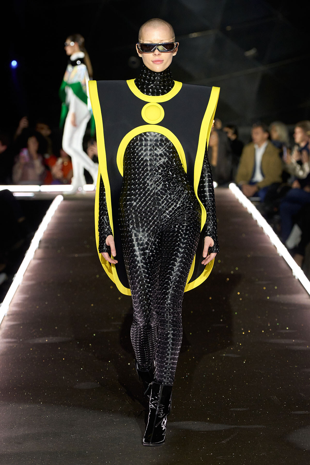 Model walks the runway in a black textured bodysuit with a futuristic neon-yellow cape-like harness and oversized sunglasses.