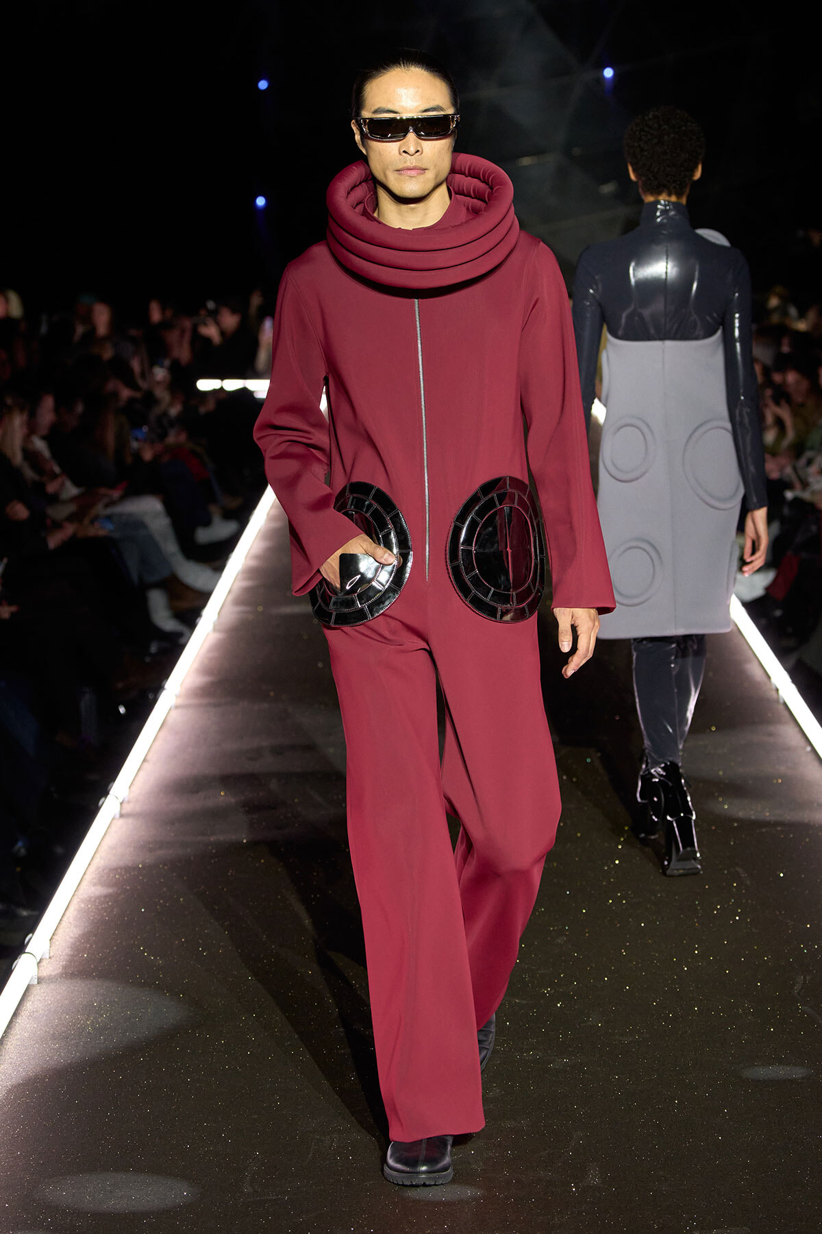 Model walks a runway wearing a burgundy avant-garde jumpsuit with oversized circular black pockets and futuristic sunglasses.