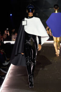 Model walking a fashion runway in a glossy black latex bodysuit with a white cape and futuristic visor helmet.