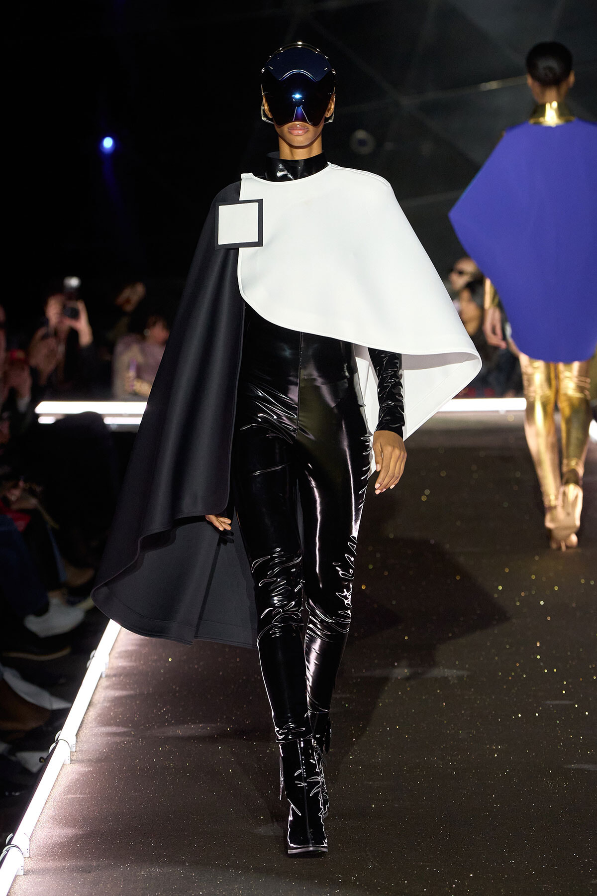 Model walking a fashion runway in a glossy black latex bodysuit with a white cape and futuristic visor helmet.