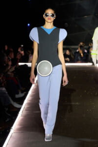 Model on a runway in a color-blocked blue and gray outfit with sculptural shoulder accents and a round metallic bag at the hip.