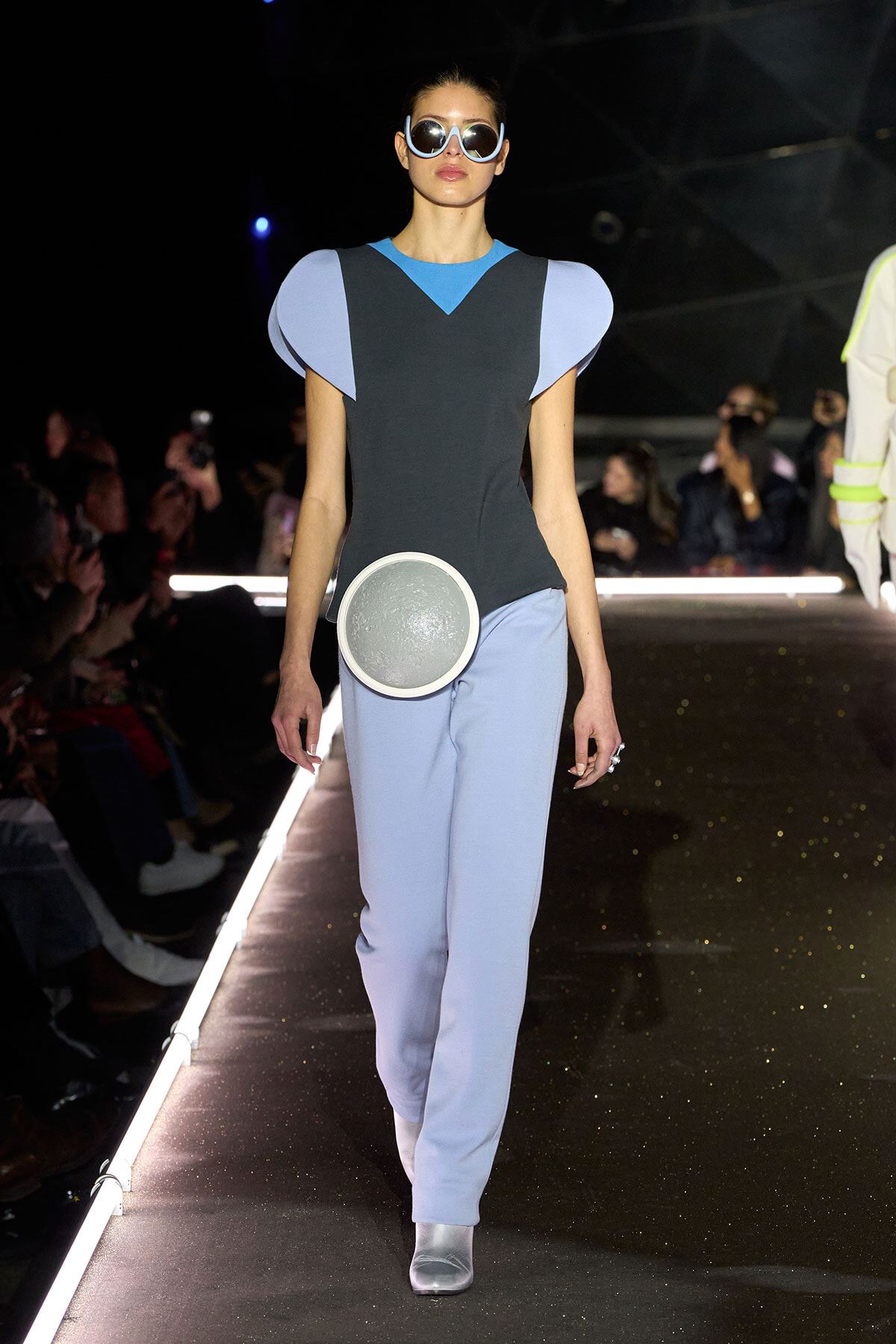 Model on a runway in a color-blocked blue and gray outfit with sculptural shoulder accents and a round metallic bag at the hip.