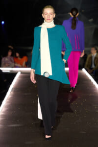 Model walking the runway in a teal tunic with a long white scarf and black pants, accessorized by a silver ball at the hip.