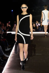 Model walks a runway in a black sleeveless dress with white curved stripes, white sunglasses, and black ankle boots while carrying a black handbag.