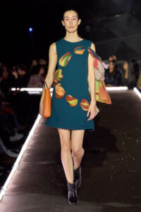Model walking a runway in a teal A-line dress with multicolored petal appliqués, black ankle boots, and an orange tote bag.