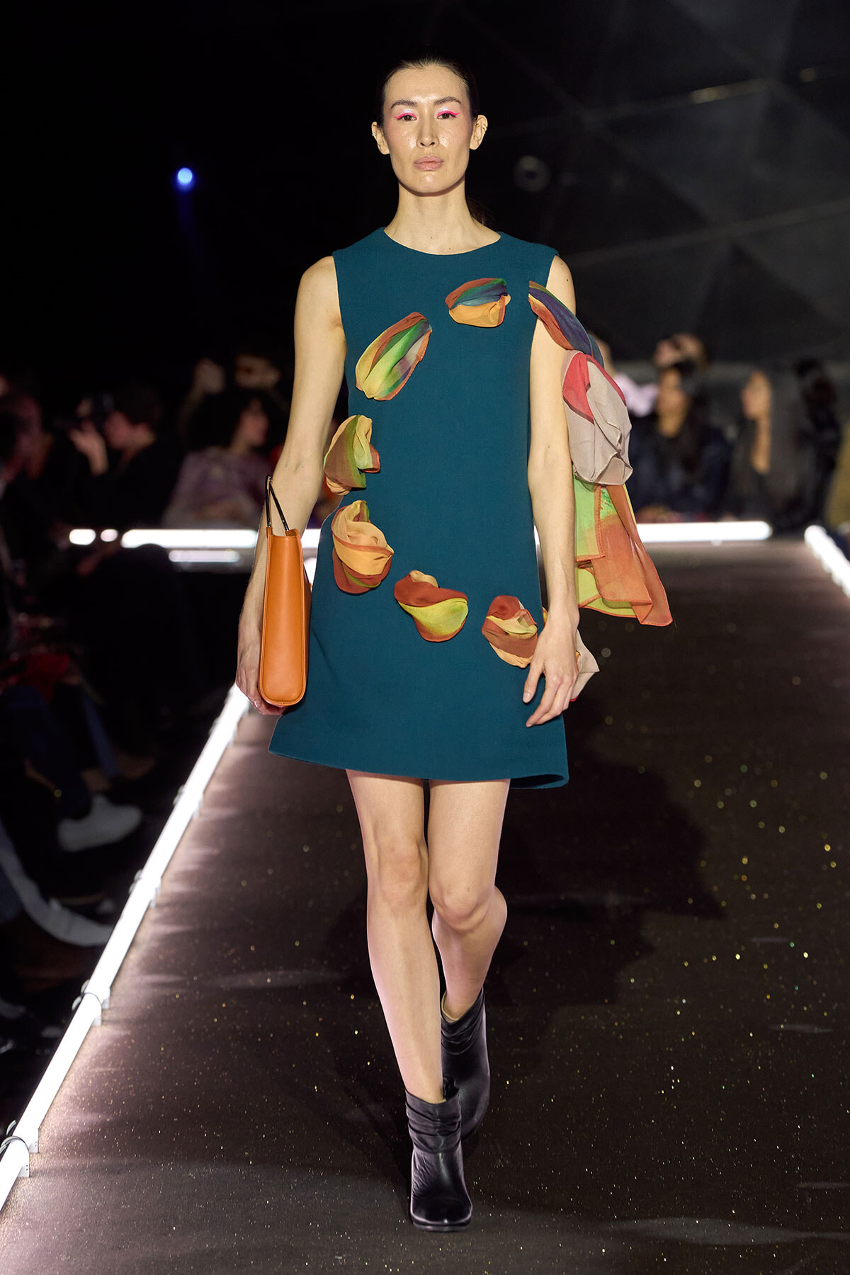 Model walking a runway in a teal A-line dress with multicolored petal appliqués, black ankle boots, and an orange tote bag.