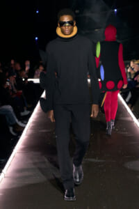 Male model walks a fashion runway in an all-black ensemble with oversized silhouette and large circular color patches attached to the sleeves and back, plus a beige high collar and futuristic sunglasses.