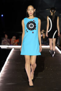 Model walks the runway in a turquoise sleeveless A-line dress with a circular black motif and colored orbs on the chest area, paired with black shoes.