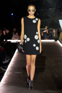 Runway model in a black sleeveless dress with silver polka dots, black ankle boots, oversized sunglasses, and a round clutch.