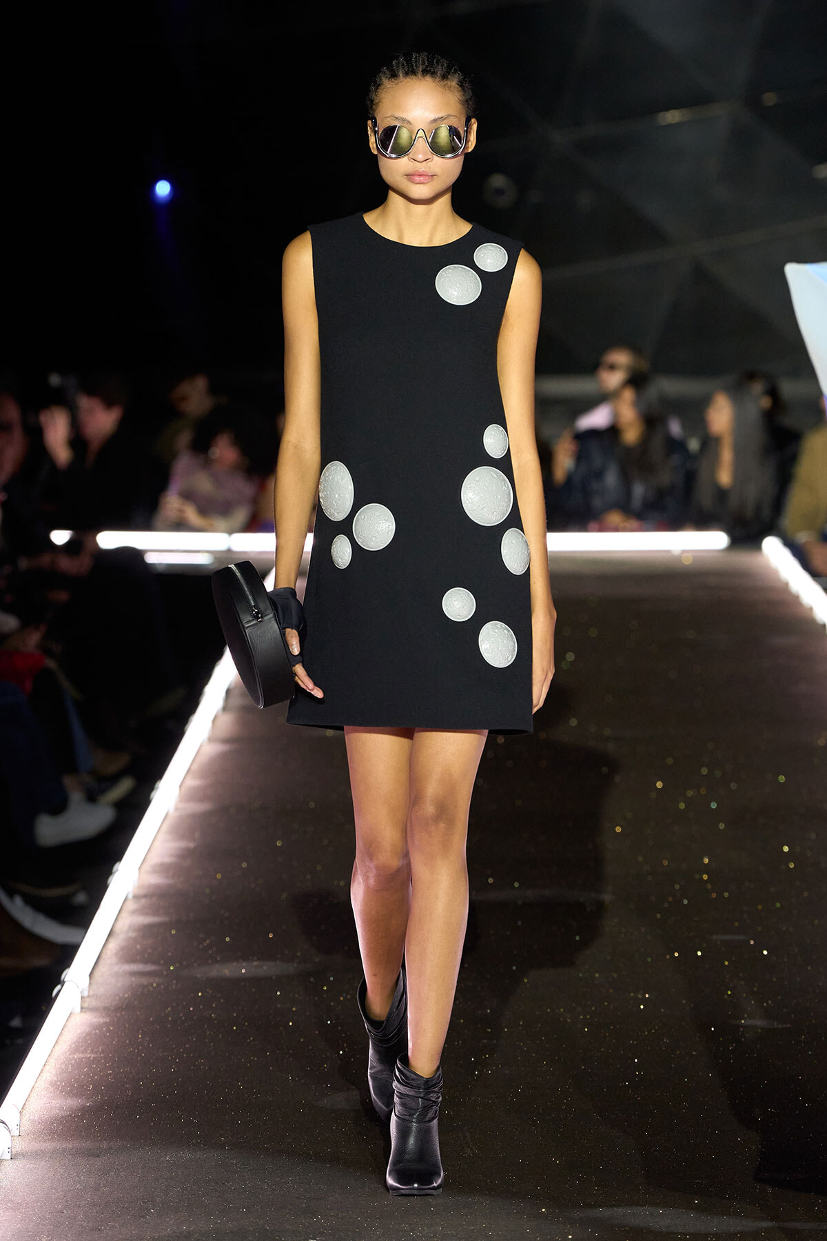 Runway model in a black sleeveless dress with silver polka dots, black ankle boots, oversized sunglasses, and a round clutch.