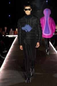 Male model on a runway wearing a black quilted padded jacket with a blue geometric panel and futuristic visor sunglasses.