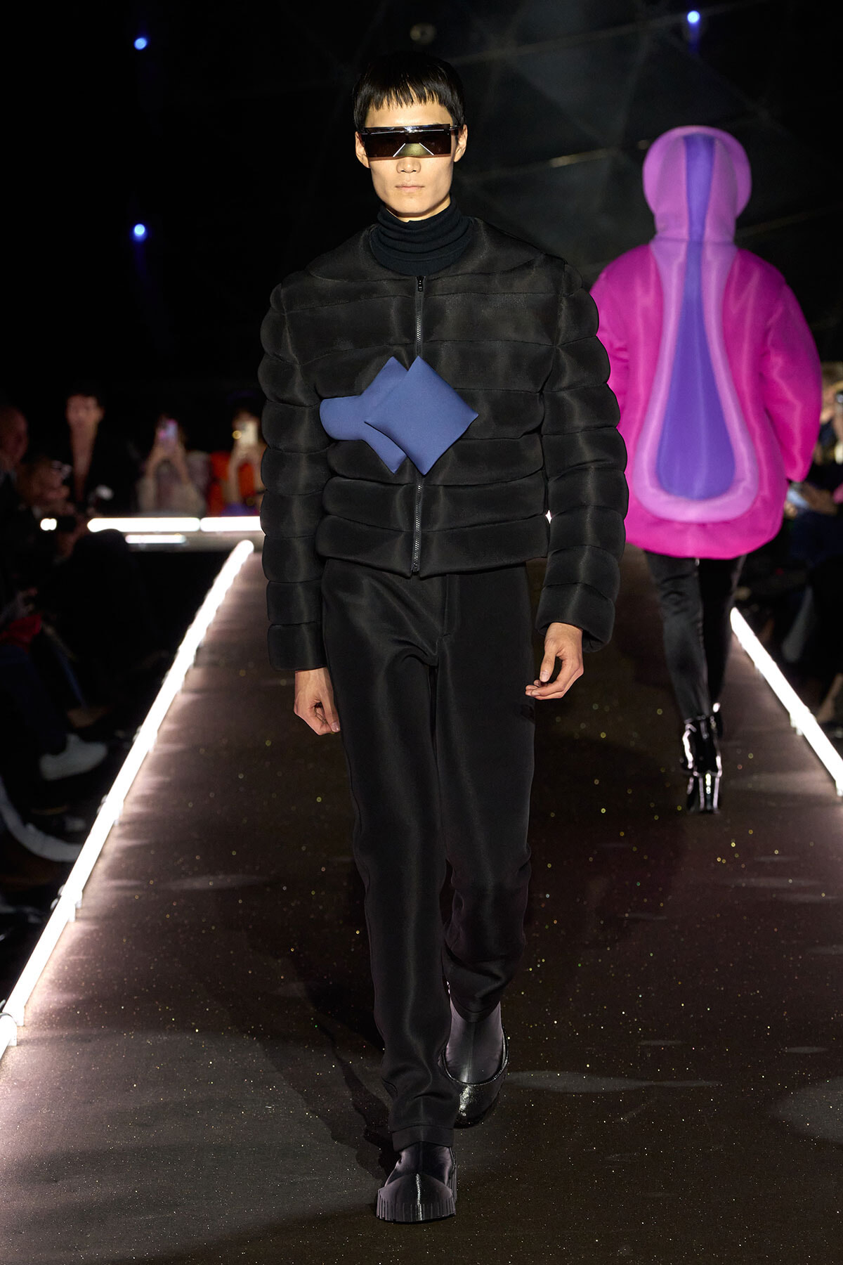 Male model on a runway wearing a black quilted padded jacket with a blue geometric panel and futuristic visor sunglasses.