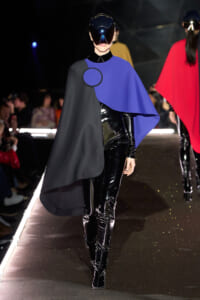 Model walking a fashion runway in a black and cobalt blue cape with circular clasp, paired with shiny black patent pants and a futuristic visor helmet.