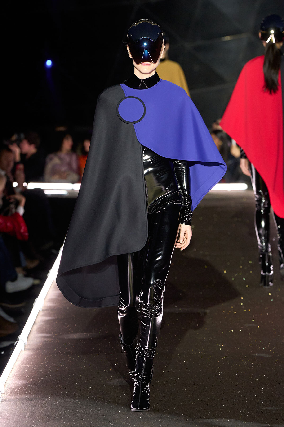 Model walking a fashion runway in a black and cobalt blue cape with circular clasp, paired with shiny black patent pants and a futuristic visor helmet.