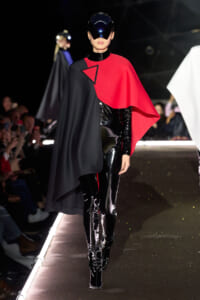 Model on a runway wearing a black and red cape over a shiny black latex bodysuit with a futuristic visor helmet.