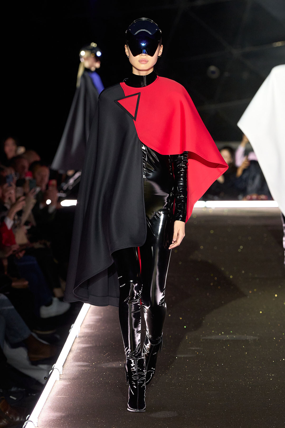 Model on a runway wearing a black and red cape over a shiny black latex bodysuit with a futuristic visor helmet.