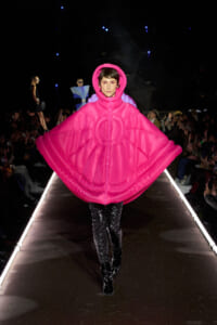 Model on a fashion show runway wearing an oversized hot-pink quilted cape with hood, paired with black sequined pants and patent boots. Audience lines the catwalk.