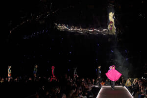 Models walk a dark runway with bright audience lights; standout pink cape outfit at the center edge of the catwalk
