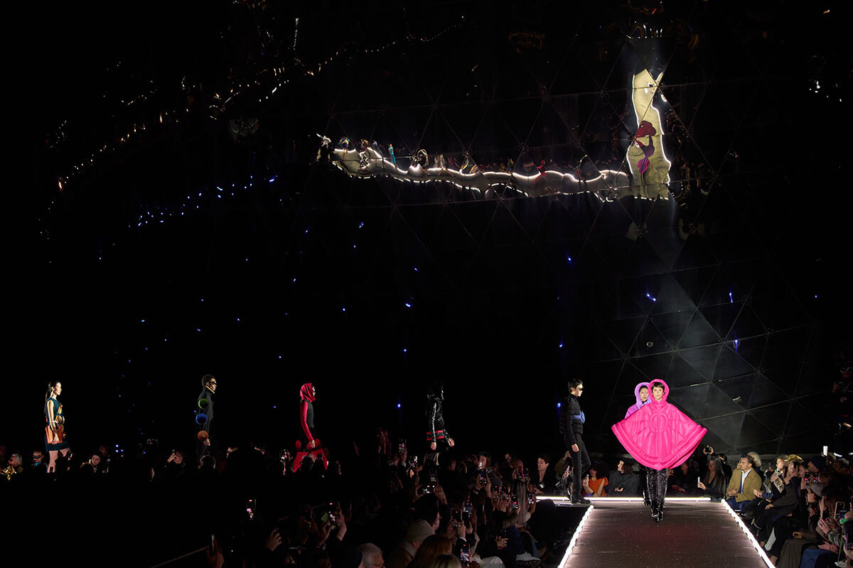 Models walk a dark runway with bright audience lights; standout pink cape outfit at the center edge of the catwalk