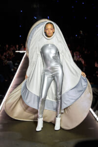 Model on a runway wearing a metallic silver jumpsuit with a large shimmering cape draped over her head and shoulders.