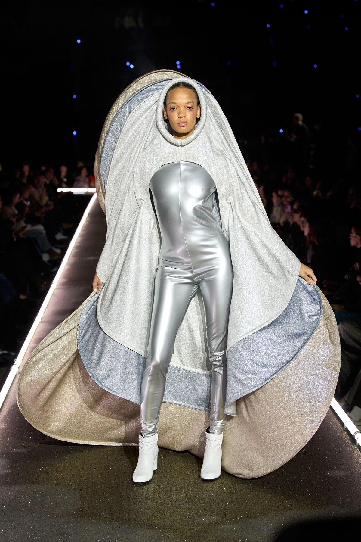 Model on a runway wearing a metallic silver jumpsuit with a large shimmering cape draped over her head and shoulders.