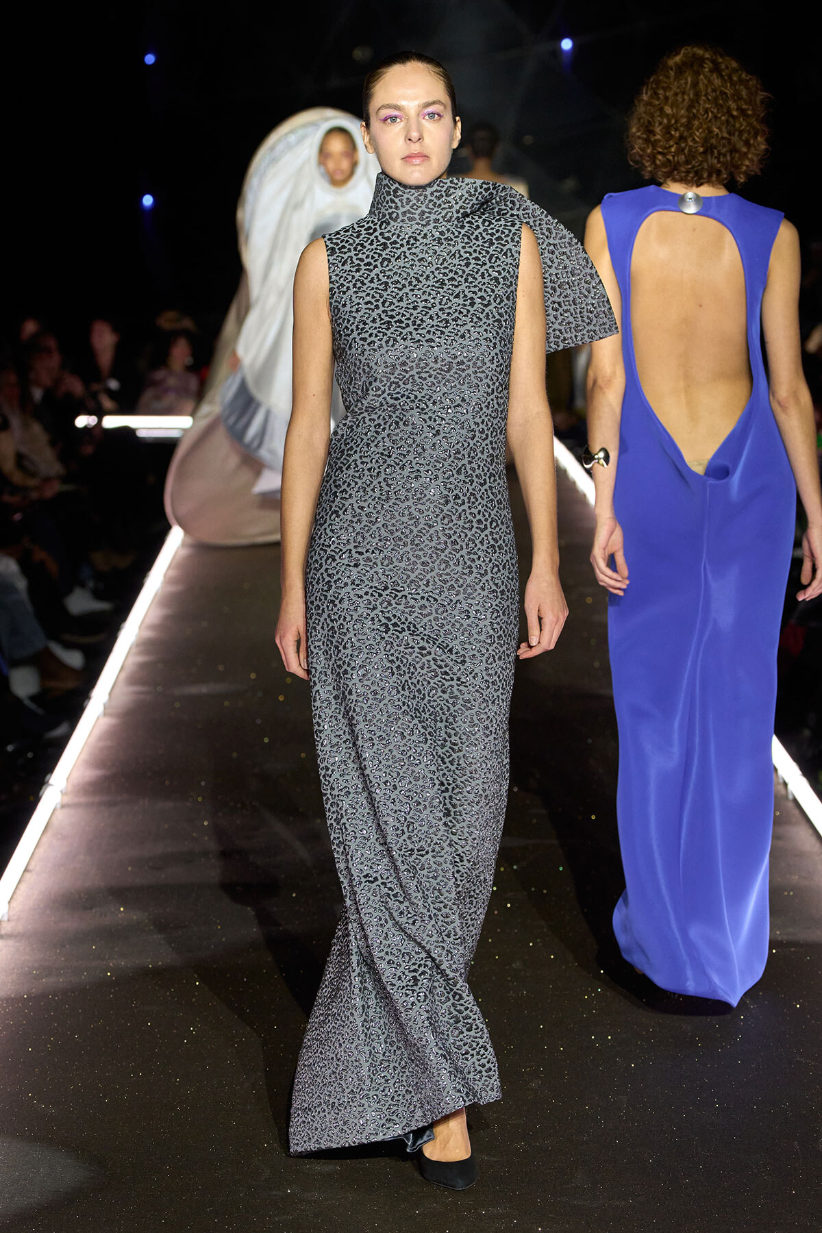 Model walks the runway in a gray leopard-print sleeveless gown with a high neckline and a draped cape detail over one shoulder.