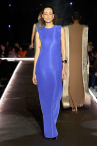 Model walks the runway in a floor-length royal blue sleeveless dress, with curly hair and pink makeup. Behind her, another model wears a beige outfit with metallic trim on a glittering catwalk edge.