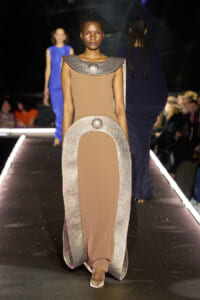 Model on a runway wearing a taupe-beige gown with a sparkling silver arch cape and metallic shoulder piece, walking the catwalk.