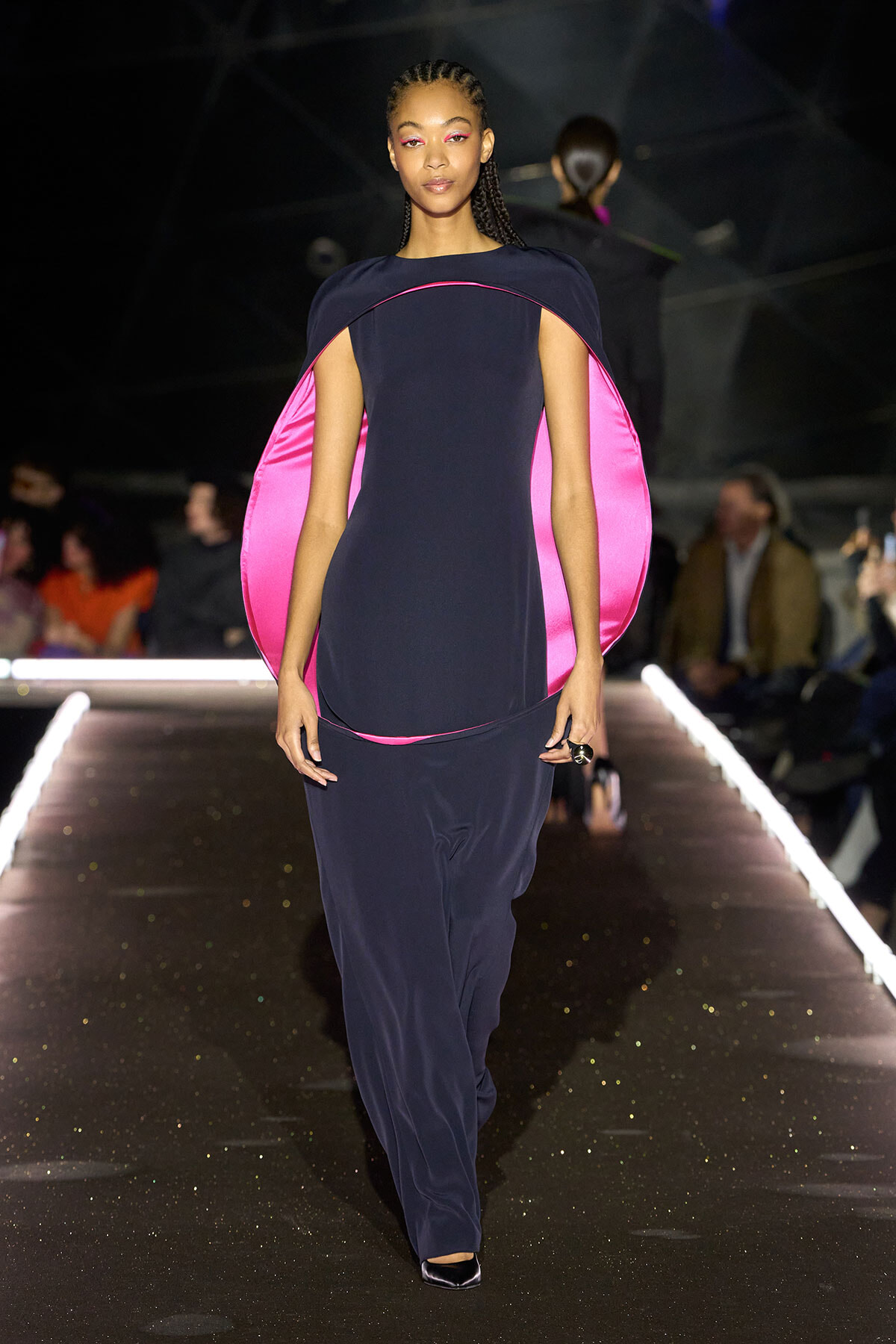 Model on a runway wearing a navy floor-length dress with a bright pink cape-like overlay draped over shoulders.