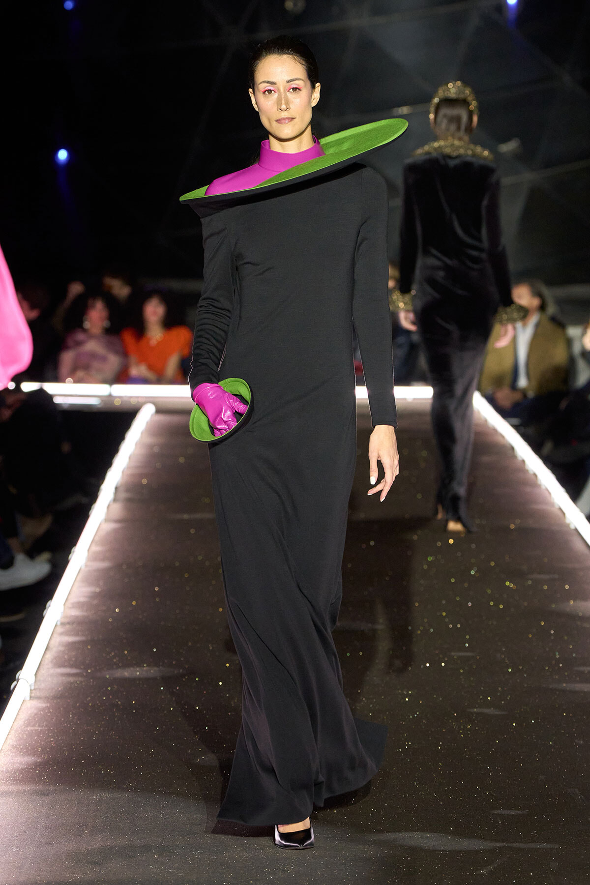 Model walks a fashion runway in a black floor-length dress with a large green halo collar, pink accents, and magenta gloves holding a green clutch.