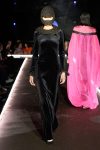 Model walks a runway in a long black velvet gown with a glittery hood and matching cuffs; pink cape outfit visible on a model in the background.