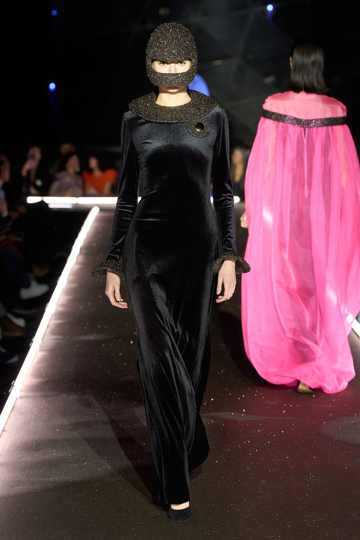 Model walks a runway in a long black velvet gown with a glittery hood and matching cuffs; pink cape outfit visible on a model in the background.