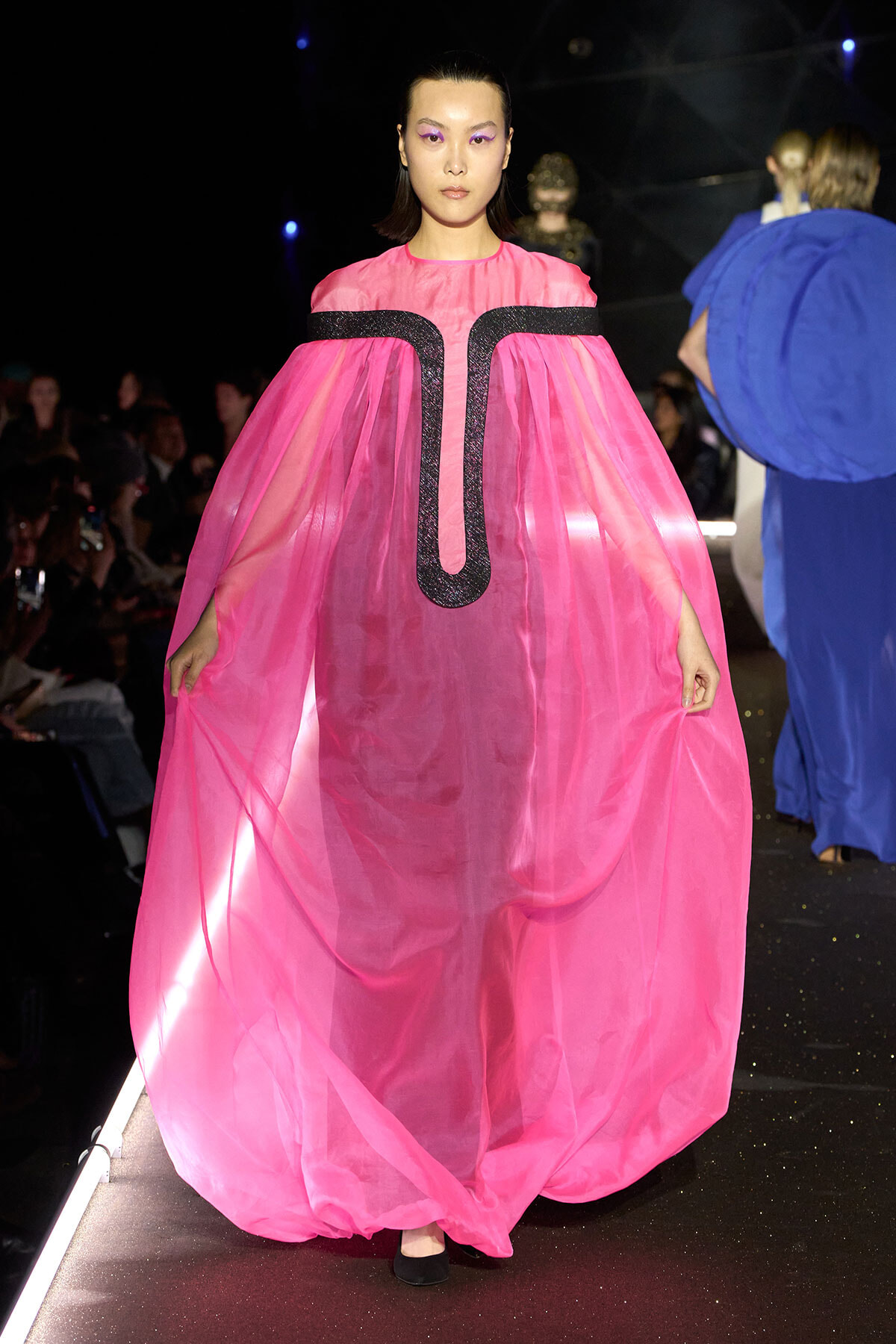 Model walking a fashion show in a bright pink, voluminous sheer cape dress with a black glitter U-shaped trim on the front surface.